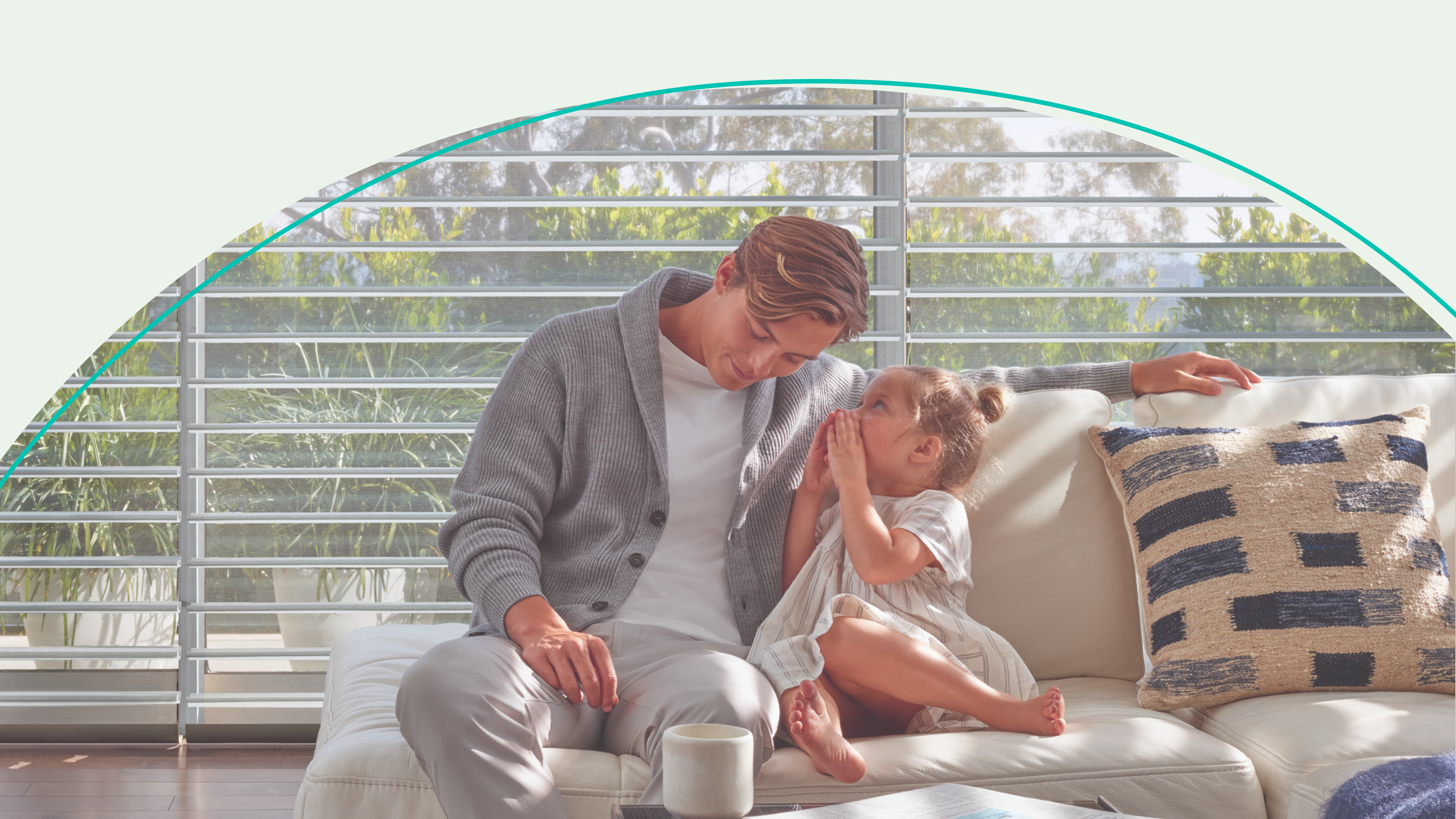 A father sits on a couch with his young daughter who is whispering in his ear. They are in front of a window.