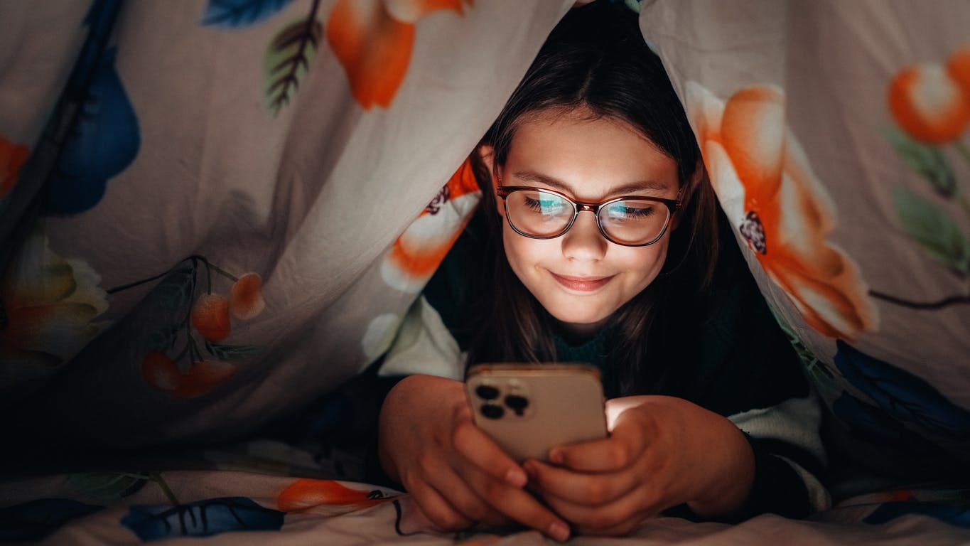 a kid under covers looking at phone