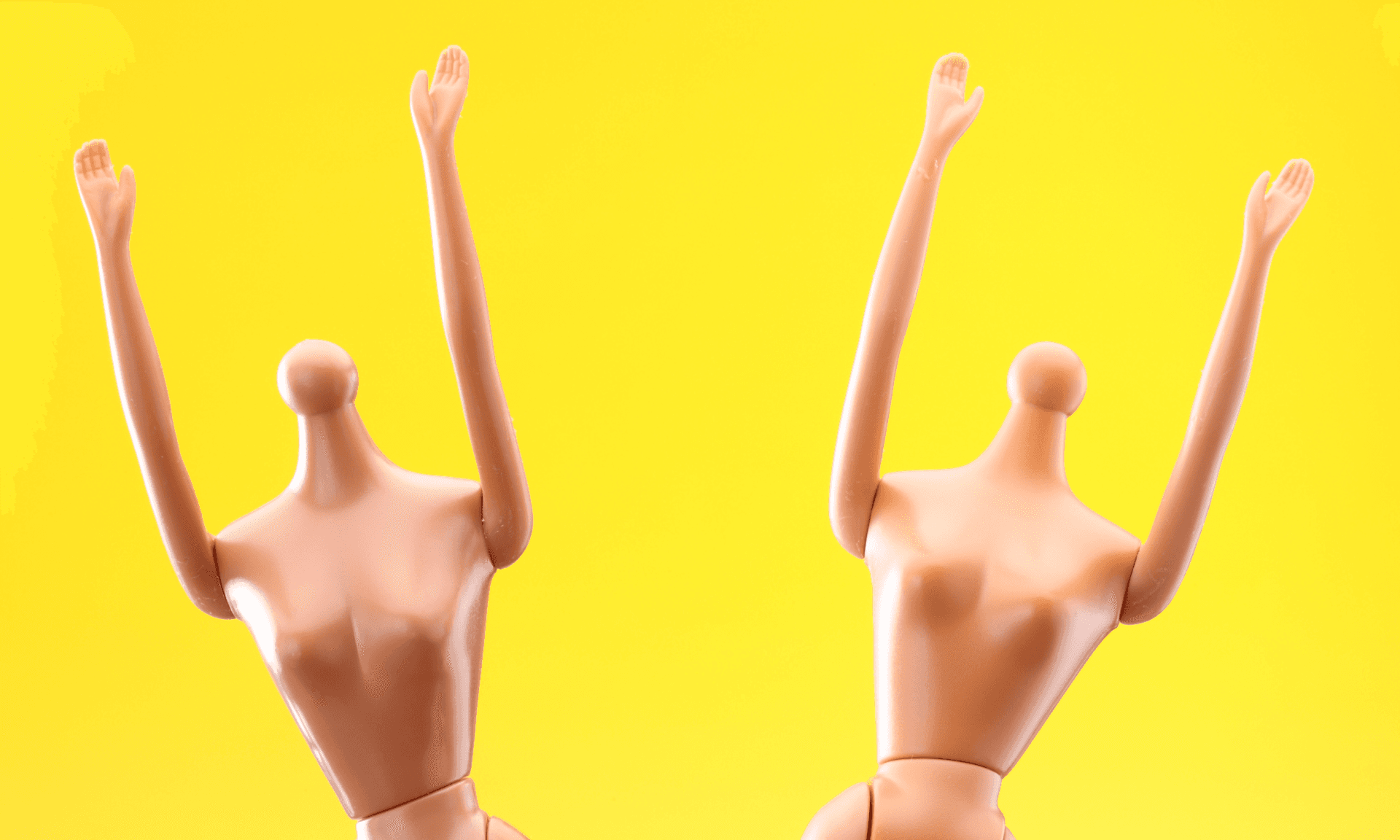 Two headless Barbie dolls with their arms in the air on a bright yellow backdrop